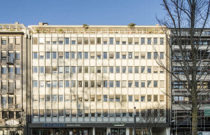 Helles Bürogebäude mit engmaschiger Rasterfassade, begrünter Dachterrasse und Gastronomie im Erdgeschoss, rechts ein eingerüsteter Nachbarbau.