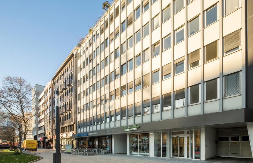 Schräge Ansicht eines Bürogebäudes mit heller Rasterfassade und verglastem Erdgeschoss, davor eine gepflasterte Fußgängerfläche.