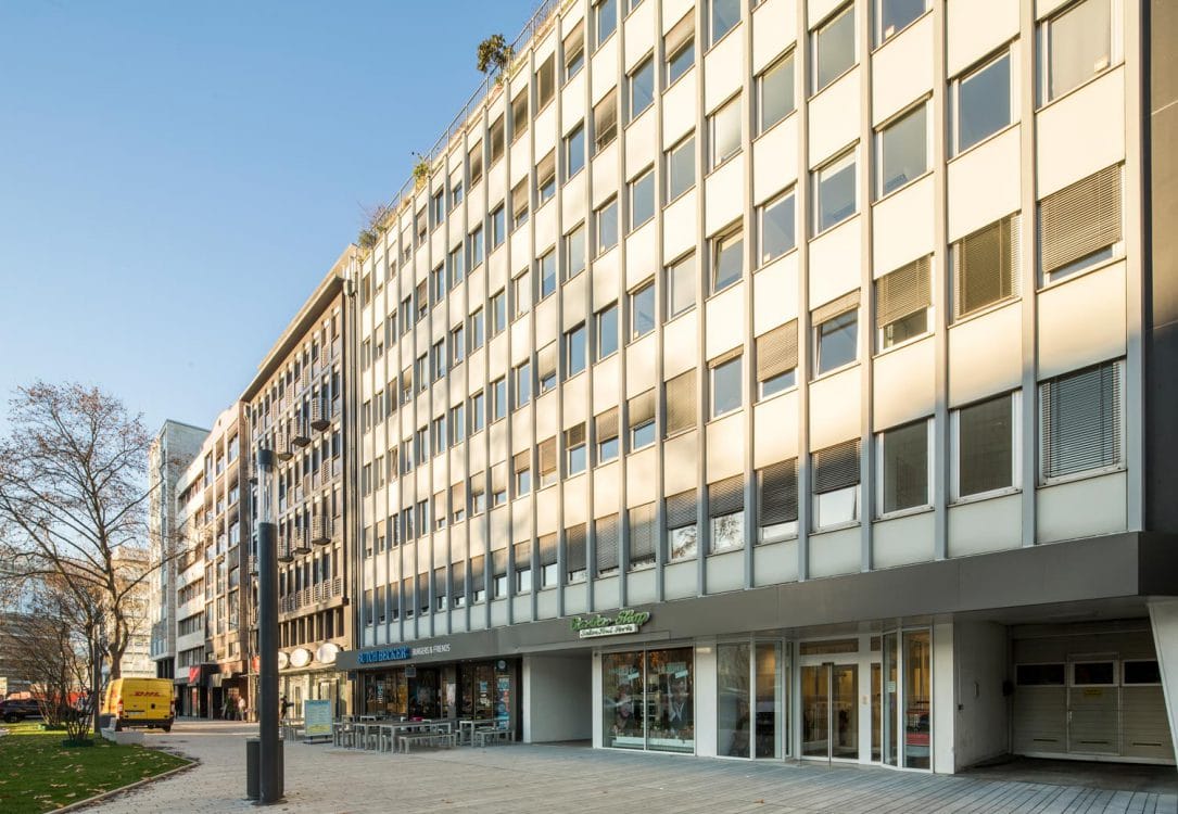 Schräge Ansicht eines Bürogebäudes mit heller Rasterfassade und verglastem Erdgeschoss, davor eine gepflasterte Fußgängerfläche.