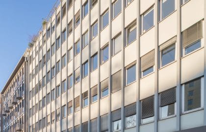 Steile Fluchtansicht einer hellen Rasterfassade über verglasten Ladeneingängen, im Vordergrund unscharfe Passanten.