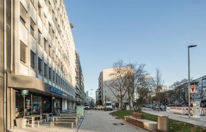 Blick entlang einer Straße mit gastronomischer Außenbestuhlung vor einem Bürogebäude, gegenüber ein Grünstreifen mit Sitzbänken.