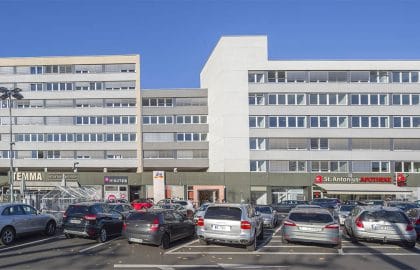 Panoramaansicht eines langgestreckten Bürogebäudes mit Ladenzeile im Erdgeschoss, davor ein voll belegter Parkplatz.