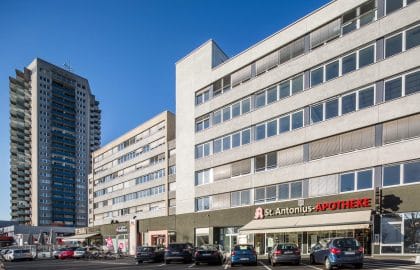 Langgestrecktes mehrgeschossiges Gebäude mit Apotheke und Ladenzeile im Erdgeschoss, davor ein Parkplatz, links ein Wohnhochhaus.