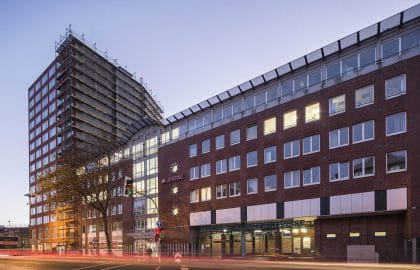 Backsteinkomplex mit Bankfiliale und Parkhauszufahrt im Erdgeschoss in der Abenddämmerung, links ein eingerüstetes Hochhaus.