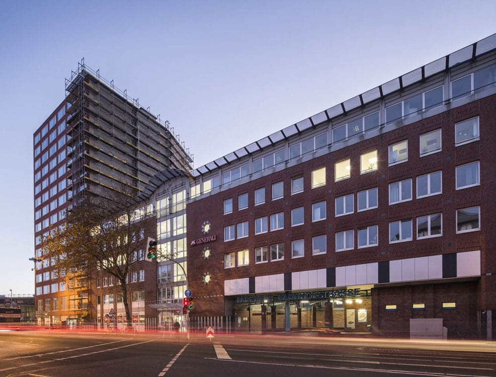 Backsteinkomplex mit Bankfiliale und Parkhauszufahrt im Erdgeschoss in der Abenddämmerung, links ein eingerüstetes Hochhaus.