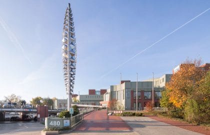 Hohe metallene Stelenskulptur auf einem gepflasterten Vorplatz, rechts ein Bürogebäude und eine Stele mit der Hausnummer 480.