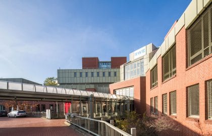 Bürogebäude aus Backstein und Metallpaneelen mit einem tonnenförmig überdachten Verbindungsgang und gepflasterter Zufahrt.