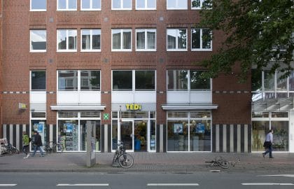 Backsteinfassade mit großen Schaufenstern und einem Einzelhandelsgeschäft im Erdgeschoss, davor Gehweg mit Passanten und Fahrrädern.