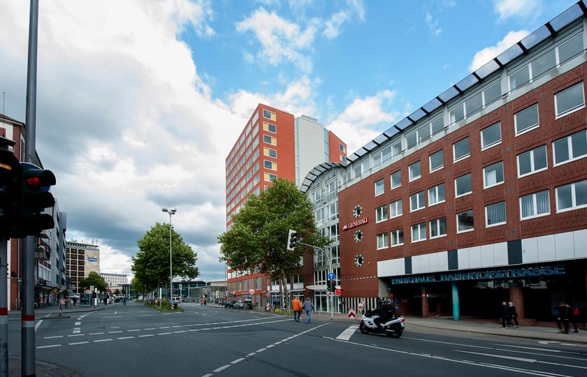 Straßenkreuzung unter Wolkenhimmel, rechts ein Backsteingebäude mit Parkhauszufahrt, dahinter ein rotes Hochhaus mit farbigen Fensterfeldern.