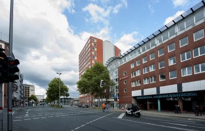 Straßenkreuzung unter Wolkenhimmel, rechts ein Backsteingebäude mit Parkhauszufahrt, dahinter ein rotes Hochhaus mit farbigen Fensterfeldern.