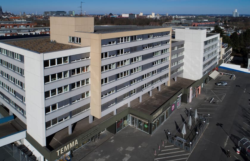 Luftaufnahme eines langgestreckten Gebäudes mit begrüntem Flachdach und Ladenzeile im Erdgeschoss, davor ein weiter Parkplatz.