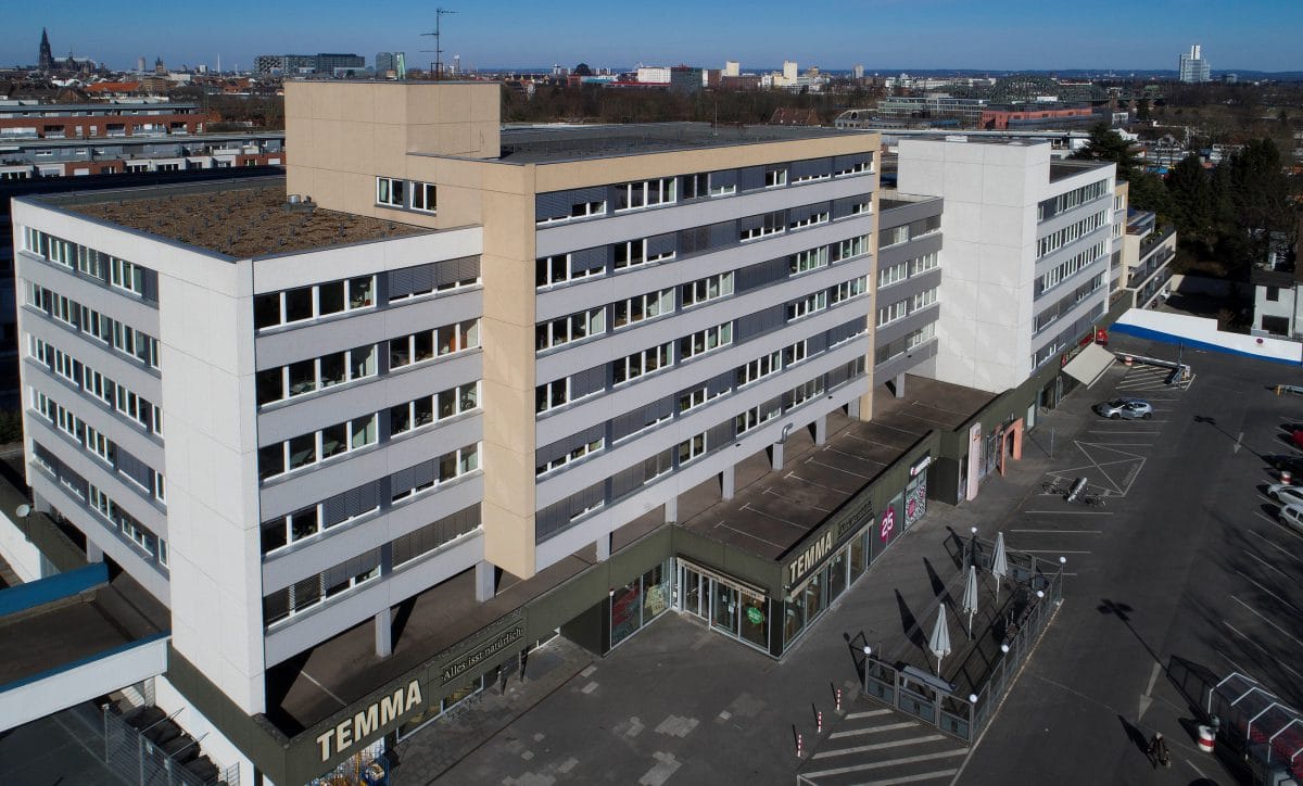 Luftaufnahme eines langgestreckten Gebäudes mit begrüntem Flachdach und Ladenzeile im Erdgeschoss, davor ein weiter Parkplatz.