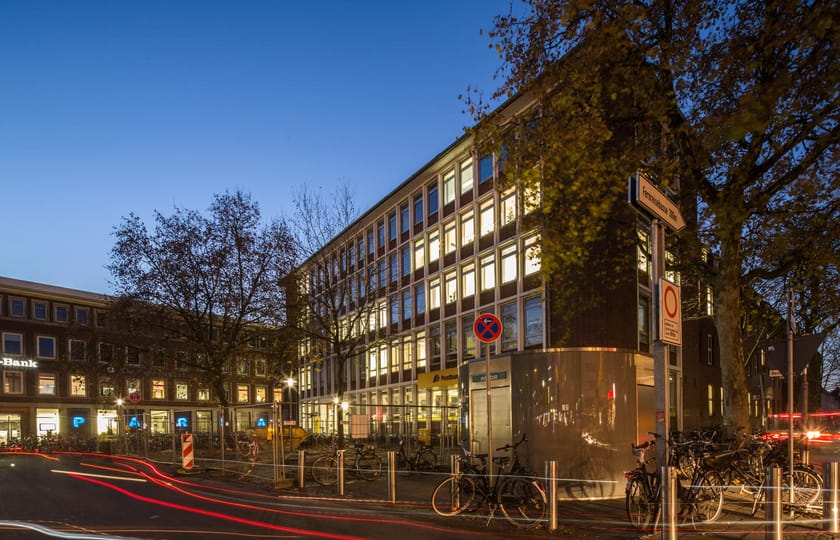 Beleuchtetes Bürogebäude mit heller Rasterfassade in der Abenddämmerung, Bankfiliale im Erdgeschoss, davor Fahrräder und Lichtspuren.
