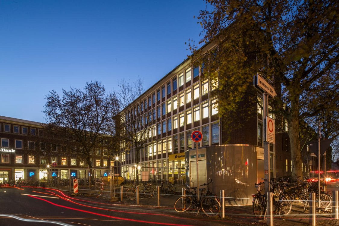 Beleuchtetes Bürogebäude mit heller Rasterfassade in der Abenddämmerung, Bankfiliale im Erdgeschoss, davor Fahrräder und Lichtspuren.