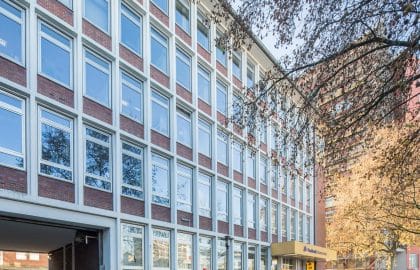 Fluchtansicht einer Fassade aus Backsteinbrüstungen und großen Fenstern, im Erdgeschoss der Eingang einer Bankfiliale.