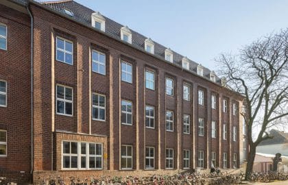 Älteres Backsteingebäude mit Schieferdach und Gauben, hohen Sprossenfenstern und einer langen Fahrradreihe entlang der Fassade.