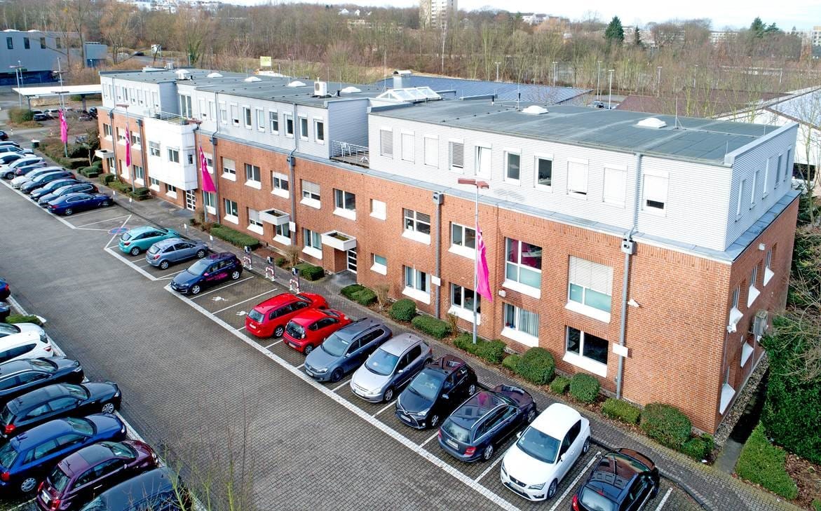 Aerial view of a two-storey brick office block with a pale set-back upper level, a car park with advertising flags in front.