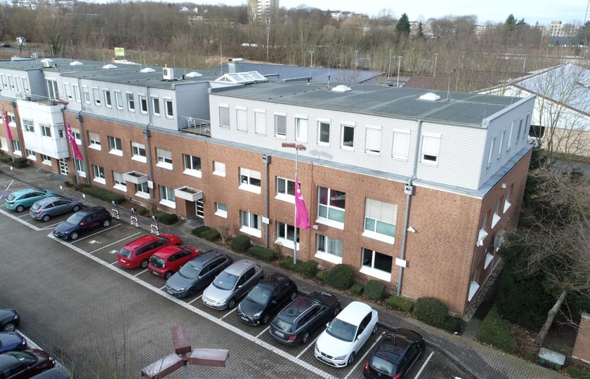 Drohnenaufnahme eines langgestreckten Bürogebäudes aus Backstein mit hellem Obergeschoss und vorgelagerten Stellplatzreihen.