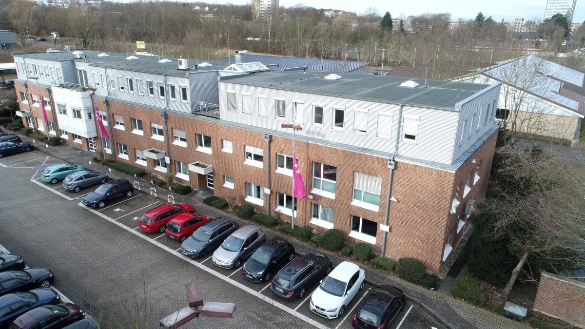 Drohnenaufnahme eines langgestreckten Bürogebäudes aus Backstein mit hellem Obergeschoss und vorgelagerten Stellplatzreihen.
