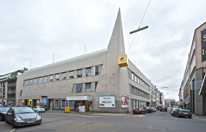 Eckgebäude mit heller Natursteinfassade und spitz zulaufender, fensterloser Gebäudekante, im Erdgeschoss Ladenlokale.