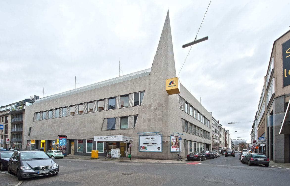 Eckgebäude mit heller Natursteinfassade und spitz zulaufender, fensterloser Gebäudekante, im Erdgeschoss Ladenlokale.