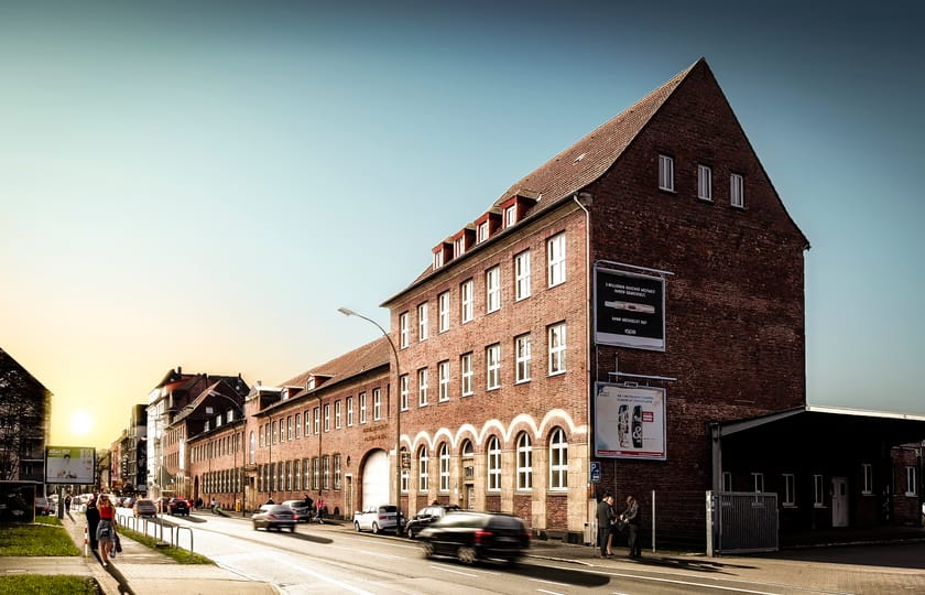 Backsteingebäude mit steilem Giebel und Rundbogenfenstern im Erdgeschoss, daran anschließend ein langgestreckter Hallentrakt im Abendlicht.