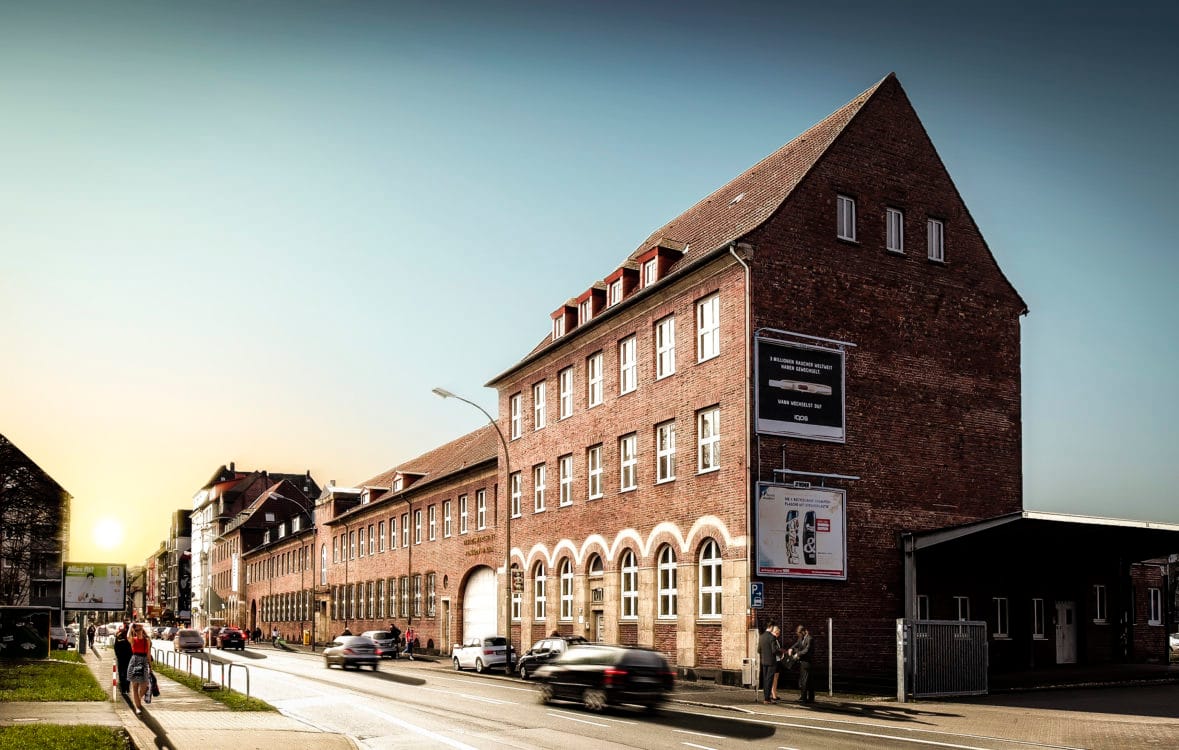 Backsteingebäude mit steilem Giebel und Rundbogenfenstern im Erdgeschoss, daran anschließend ein langgestreckter Hallentrakt im Abendlicht.