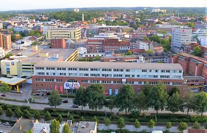 Luftaufnahme über eine Stadt mit einem langgestreckten Backsteingebäude mit hellem Aufbau im Vordergrund, dahinter dichte Bebauung.