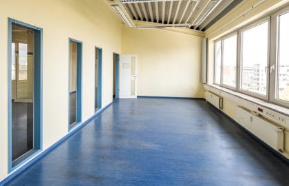 Leerer Büroraum mit gelben Wänden, blauem Bodenbelag, blau gefassten Türlaibungen und einem durchgehenden Fensterband.