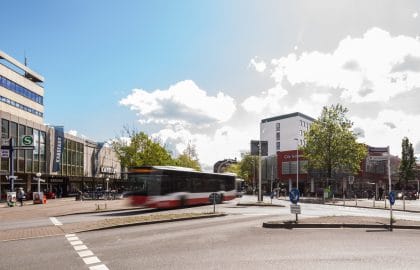 Innerstädtische Straßenkreuzung mit einem vorbeifahrenden Bus, links ein Kaufhaus mit Bahnzugang, rechts ein moderner Geschäftsbau.