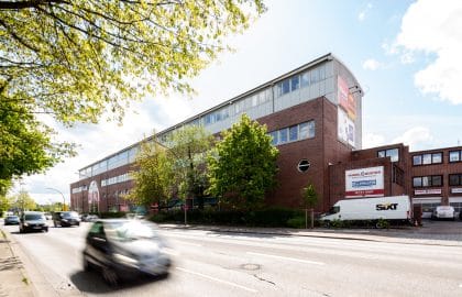 Langgestrecktes Backsteingebäude mit verglastem Staffelgeschoss an einer mehrspurigen Straße, davor Bäume und fahrende Autos.
