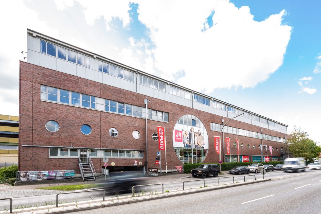 Backsteinfassade mit runden Fenstern, einem großen Kreisfenster und verglastem Obergeschoss, davor eine mehrspurige Straße.
