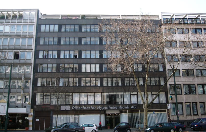 Dunkel verkleidetes Bürogebäude mit durchgehenden Fensterbändern und Bankfiliale im Erdgeschoss, eingefasst von helleren Nachbarhäusern.