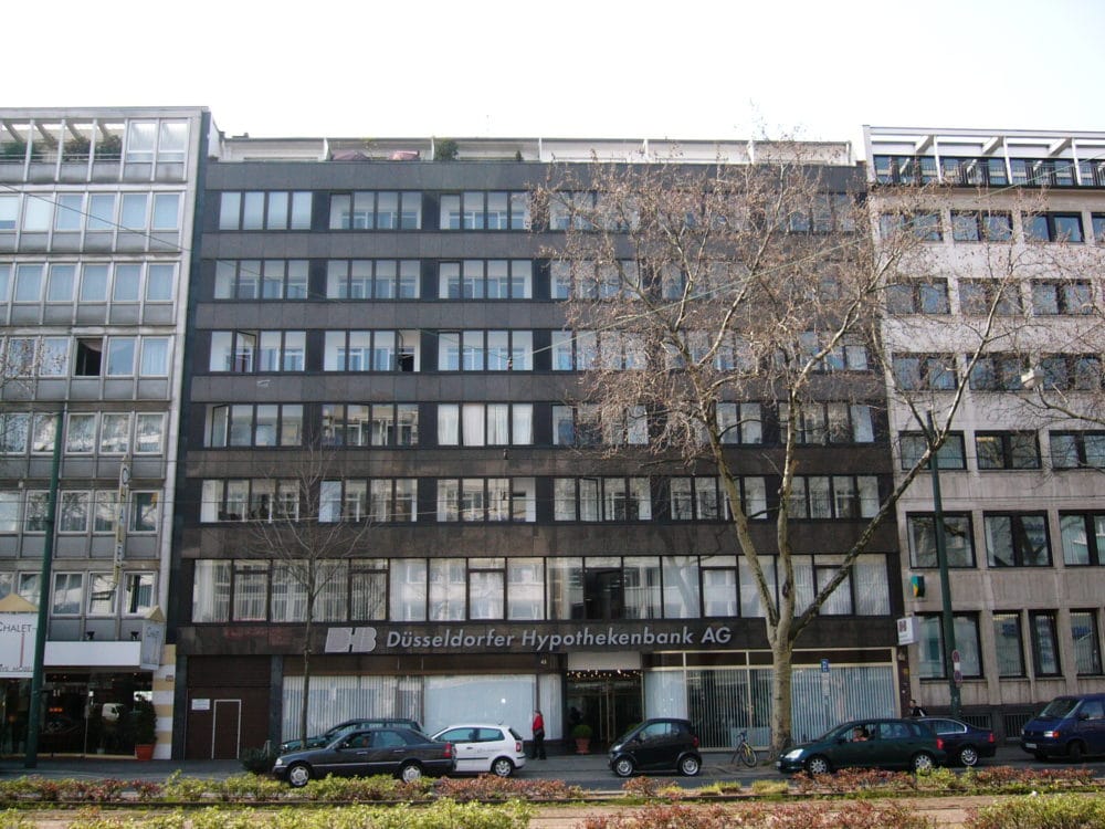 Dunkel verkleidetes Bürogebäude mit durchgehenden Fensterbändern und Bankfiliale im Erdgeschoss, eingefasst von helleren Nachbarhäusern.