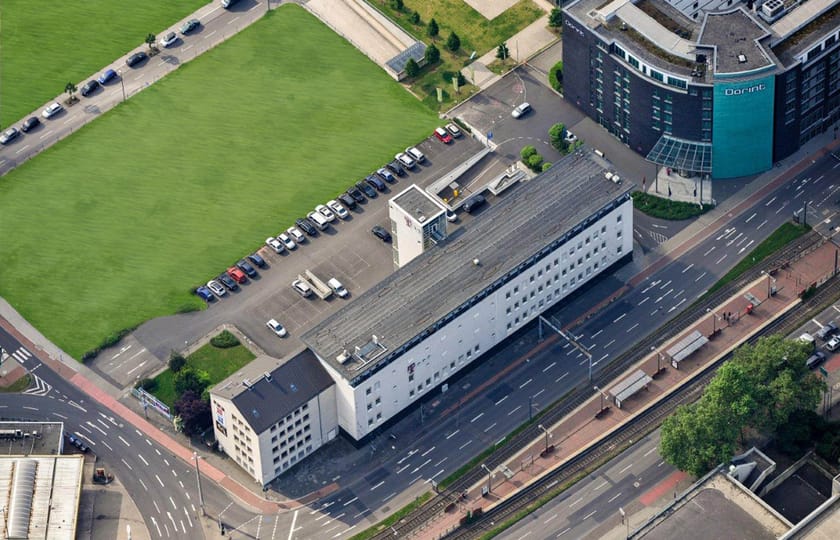 Schräges Luftbild eines langgestreckten weißen Gebäudes an einer mehrspurigen Straße mit Bahngleisen, links eine große Rasenfläche.