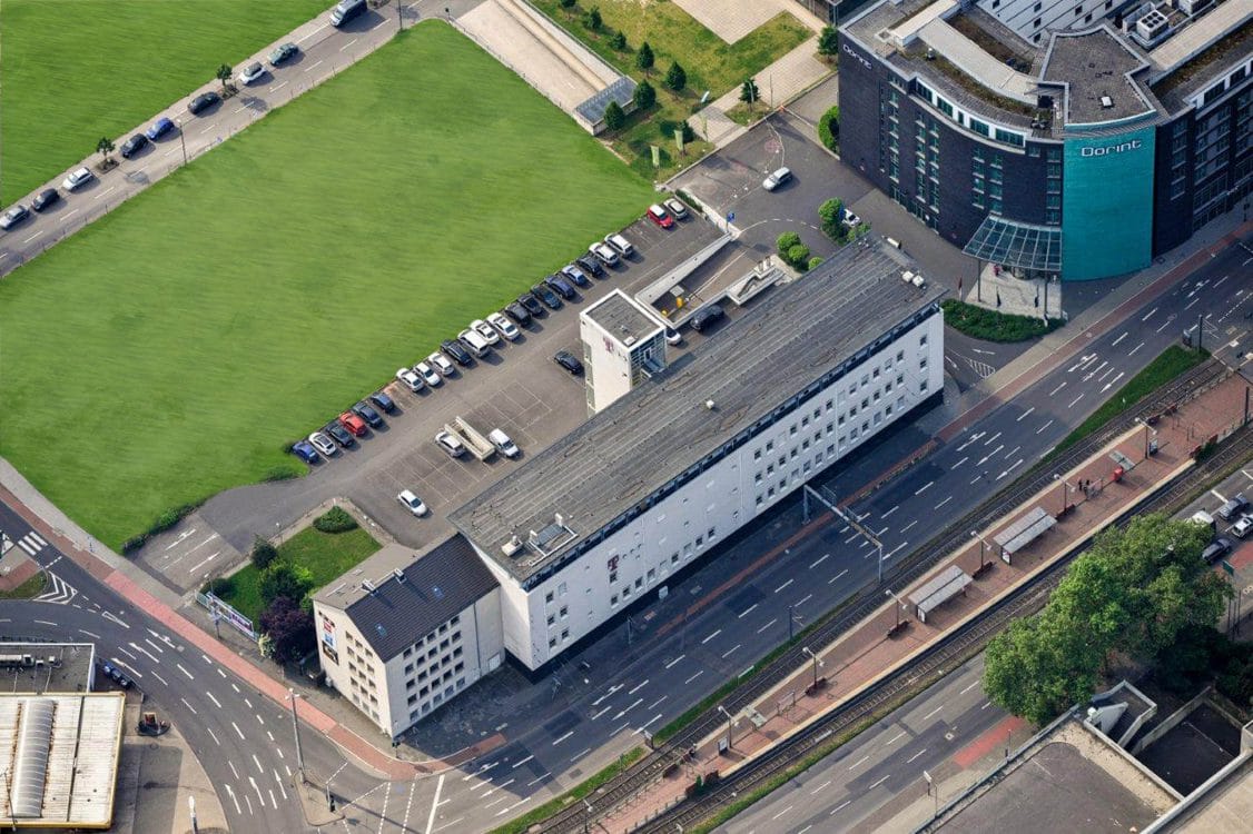 Schräges Luftbild eines langgestreckten weißen Gebäudes an einer mehrspurigen Straße mit Bahngleisen, links eine große Rasenfläche.