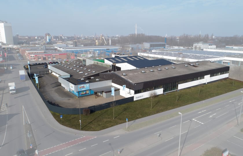 Drohnenaufnahme eines Gewerbeobjekts mit flachen Hallen, weiß-dunkler Fassade und Hoffläche, umgeben von einem Gewerbegebiet.