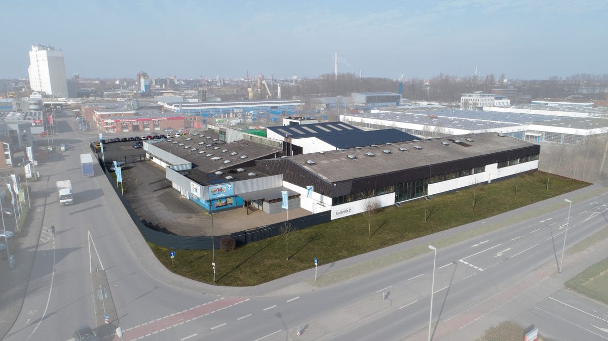 Drohnenaufnahme eines Gewerbeobjekts mit flachen Hallen, weiß-dunkler Fassade und Hoffläche, umgeben von einem Gewerbegebiet.