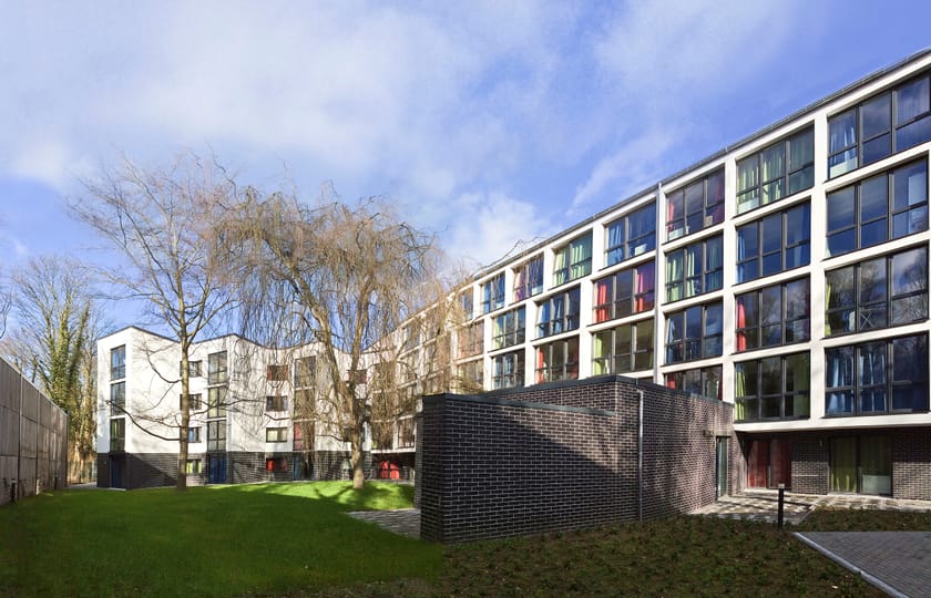 Mehrgeschossige Wohnanlage mit weißer Fassade und farbig gefassten Fensterlaibungen, davor eine Rasenfläche mit kahlem Baum.