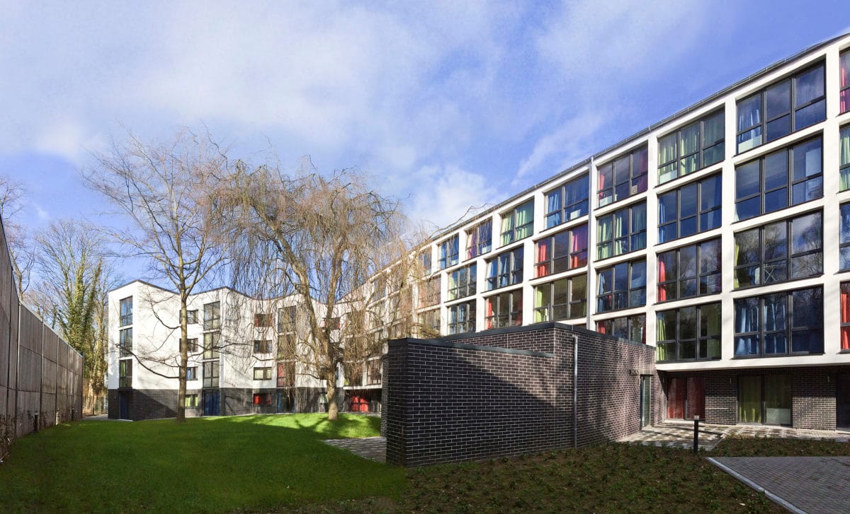 Mehrgeschossige Wohnanlage mit weißer Fassade und farbig gefassten Fensterlaibungen, davor eine Rasenfläche mit kahlem Baum.