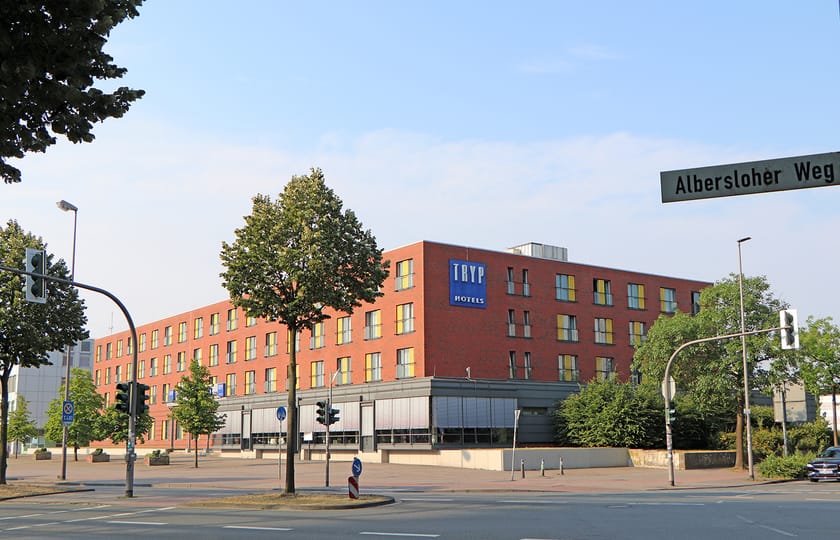 Mehrgeschossiger Hotelbau aus rotem Backstein mit gelb gefassten Fensterlaibungen und Hotelschild, an einer begrünten Straßenkreuzung.