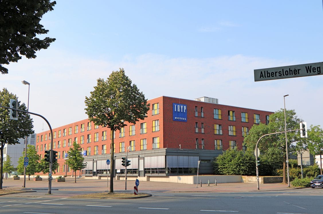 Mehrgeschossiger Hotelbau aus rotem Backstein mit gelb gefassten Fensterlaibungen und Hotelschild, an einer begrünten Straßenkreuzung.