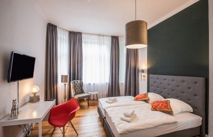 Hotelzimmer mit Doppelbett vor dunkelgrüner Wand, Erkerfenster mit Vorhängen, Schreibtisch mit rotem Schalenstuhl und Dielenboden.