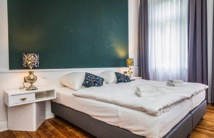 Frisch bezogenes Doppelbett vor einer dunkelgrünen Wandfläche, flankiert von zwei Nachttischlampen, daneben ein Fenster mit Vorhängen.