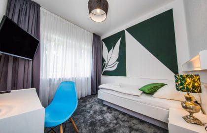 Kleines Hotelzimmer mit Einzelbett, grafischer Wandgestaltung in Grün und Weiß, Schreibtisch mit blauem Schalenstuhl und grauem Teppichboden.