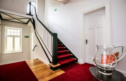 Treppenhaus mit dunkelgrün gestrichenem Holzgeländer, rotem Läufer, Stuckleiste und einer großen Glasvase auf einem Beistelltisch.