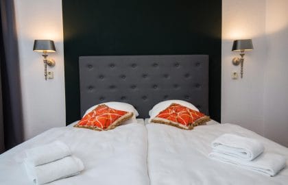 Bettkopfende mit grau gepolstertem Kopfteil vor dunkelgrüner Wand, zwei Wandleuchten, orangefarbene Kissen und gefaltete Handtücher.
