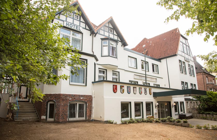 Weiß verputztes Haus mit Fachwerkgiebel, rotem Ziegeldach und Hotelschriftzug, über dem Eingang eine Reihe bunter Wappenschilder.