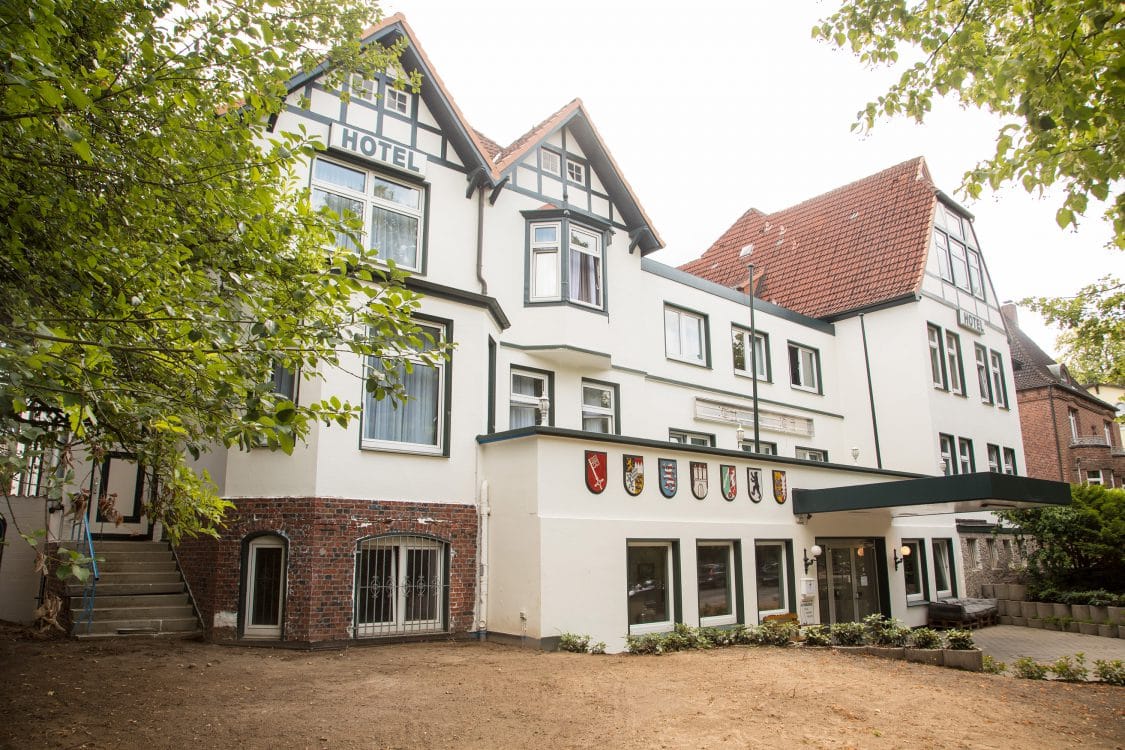 Weiß verputztes Haus mit Fachwerkgiebel, rotem Ziegeldach und Hotelschriftzug, über dem Eingang eine Reihe bunter Wappenschilder.