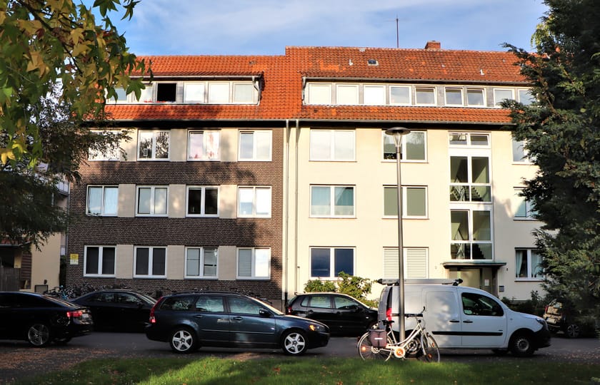 Mehrfamilienhaus mit rotem Ziegeldach, links dunkler Klinkerfassade und rechts hellem Putz, davor parkende Autos und eine Rasenfläche.